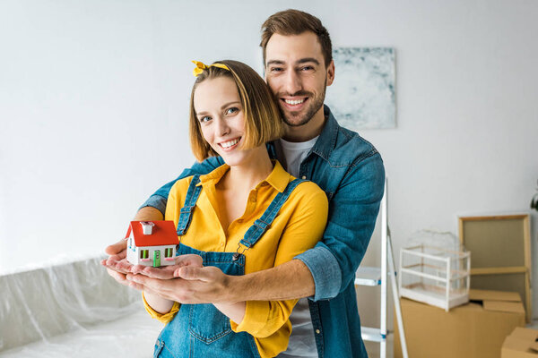 Smiling couple holding toy house and looking at camera at home