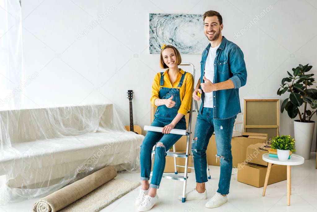 full length view of couple with cardboard boxes and blueprint showing thumbs up