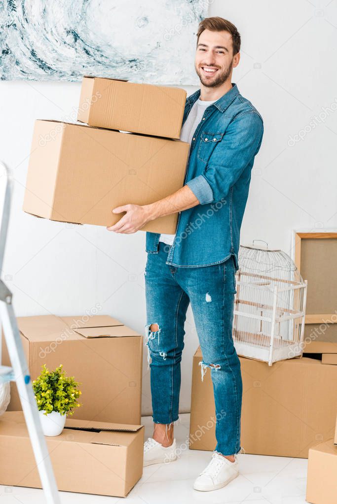 full length view of laughing man in jeans holding cardboard boxes and looking at camera