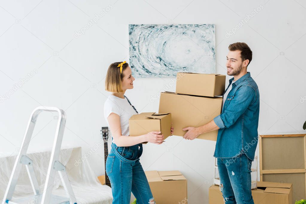 Joyful couple in jeans holding cardboard boxes and looking at each other at home