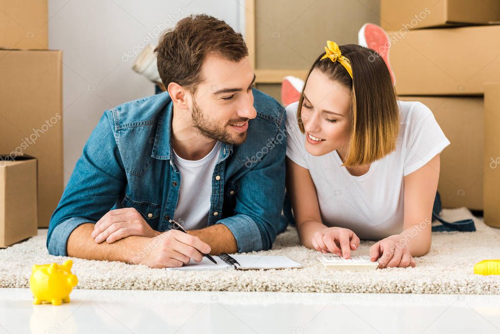 smiling couple lying on carpet with notebook, pen and calculator