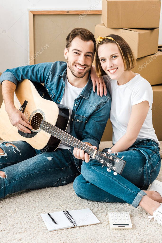 Smiling man sitting on carpet with wife and playing acoustic guitar
