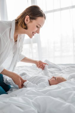 Beyaz gömlekli gülen genç anne yatakta bebeğe bakıyor