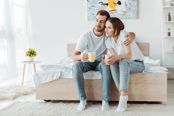 couple with cups of coffee sitting on bed and looking away