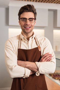 Gözlük ve kahverengi önlük içinde gülümseyen barista çapraz kolları ile kamera bakıyor