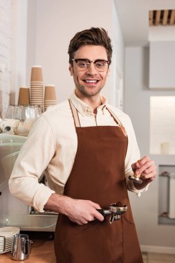 Bardak ve kahverengi önlük zemin kahve ve kurcaper ile portafilter tutan Gülümseyen barista