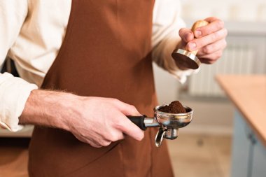 Cropped view of barista in brown apron holding portafilter with ground coffee and tamper clipart