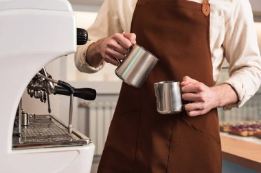 Cropped view of barista in brown apron holding steel jugs clipart