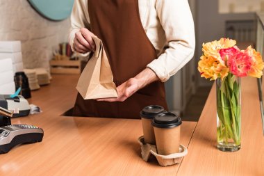 Partial view of barista in brown apron with paper bag and coffee to go clipart