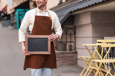 Sokakta tebeşir menüsü tutan kahverengi önlük gülümseyen barista Kırpılmış görünümü