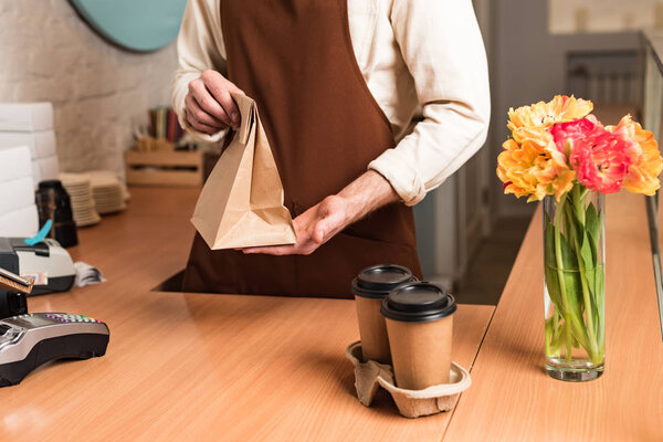 Partial view of barista in brown apron with paper bag and coffee to go