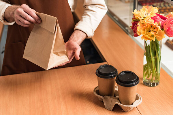 Partial view of barista in brown apron with paper bag and coffee to go