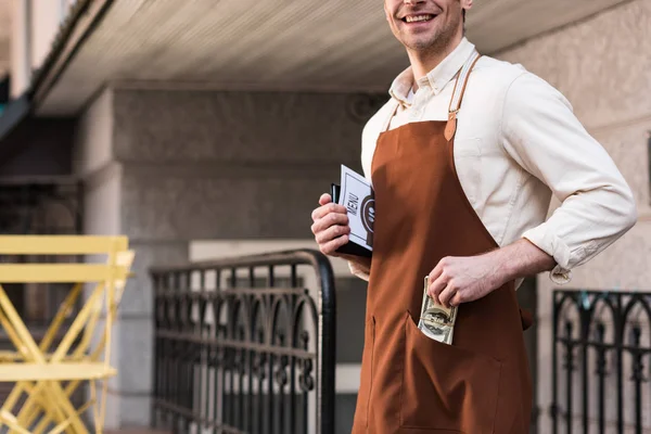 Partial view of barista in brown apron putting money in pocket
