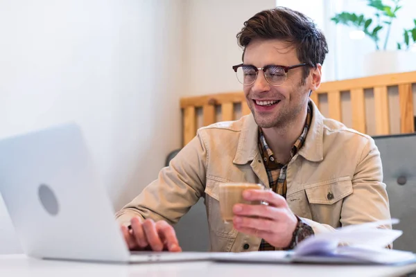 Uśmiechnięty Freelancer Okularach Trzymając Filiżankę Kawy Wpisując Klawiaturze Laptopa — Zdjęcie stockowe