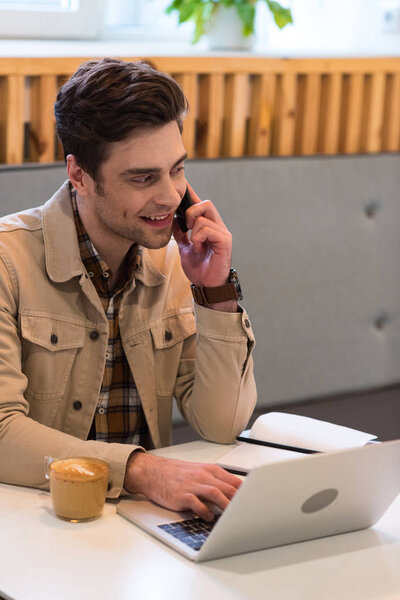Smiling freelancer in jacket talking on smartphone in cafe