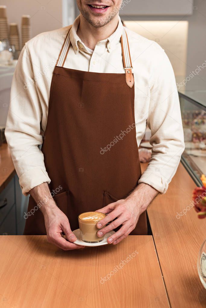 Partial view of smiling barista in brown apron holding cup of coffee