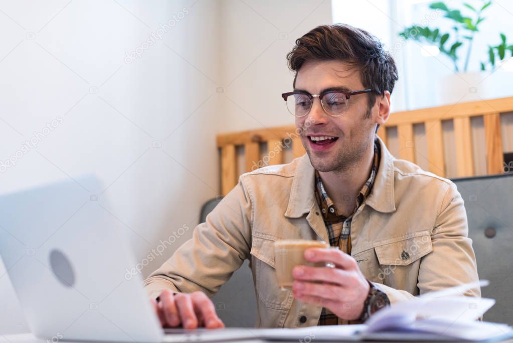 Smiling freelancer in glasses holding cup of coffee and typing on laptop keyboard