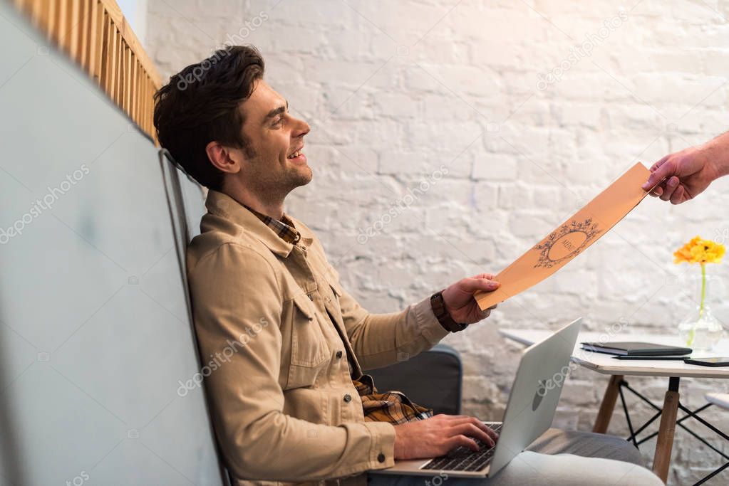 Smiling freelancer with laptop holding menu in cafe
