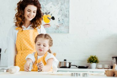 happy mother in yellow polka dot apron looking at daughter holding dough molds in kitchen clipart