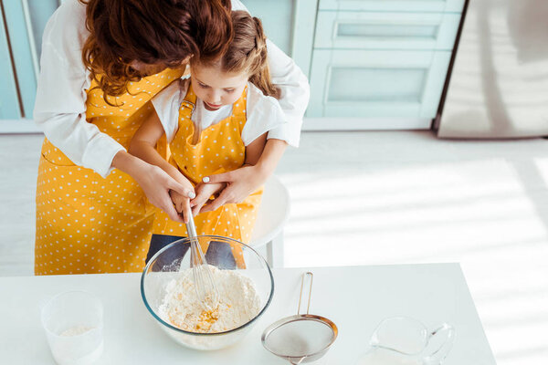 overhead view of mother and daughter in polka dot aprons mixing flour and eggs with balloon whisk in bowl