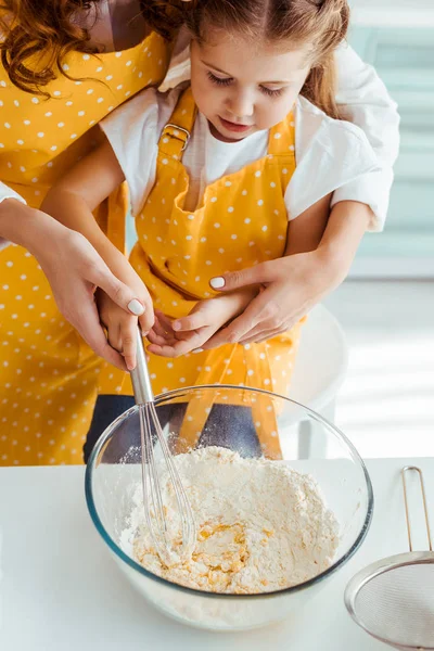 ボウルに風船の泡立て器で小麦粉と卵を混合するエプロンの母と娘 — ストック写真