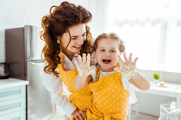 Улыбающаяся мать смотрит на смеющуюся счастливую дочь с грязными руками в тесте
