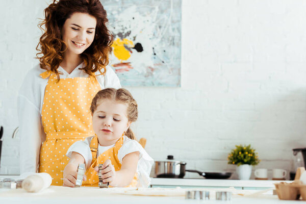 happy mother in yellow polka dot apron looking at daughter holding dough molds in kitchen