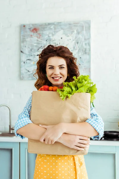 熟した果物や野菜と紙袋を抱きしめる水玉エプロンで笑顔の女性 — ストック写真