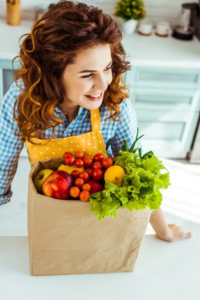 快乐的女人靠近纸袋与成熟的水果和蔬菜在厨房桌子上 — 图库照片