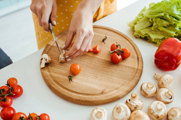 cropped view of woman in polka dot apron cutting mushrooms and cherry tomatoes on chopping board