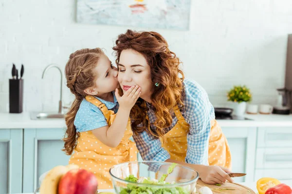 可爱的女儿在波尔卡点黄色围裙亲吻母亲与闭着眼睛在厨房 — 图库照片