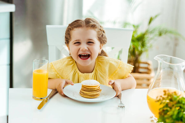 happy excited kid sitting at table, laughing and holding plate with pancakes