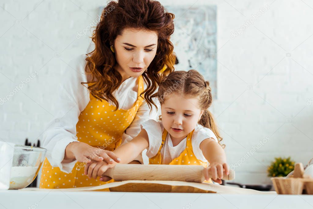 young mother helping daughter rolling out dough on baking parchment paper