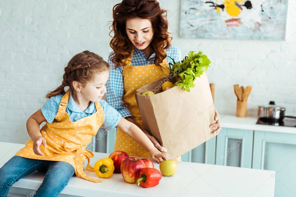 mother in polka dot apron holding paper bag while daughter sitting on table and touching apples and bell peppers