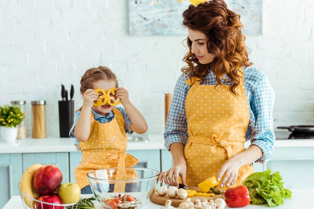 mother in polka dot yellow apron giving looking at daughter with bell pepper slices near eyes