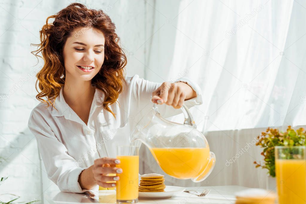 beautiful happy woman sitting at table and pouring orange juice in glass while having breakfast