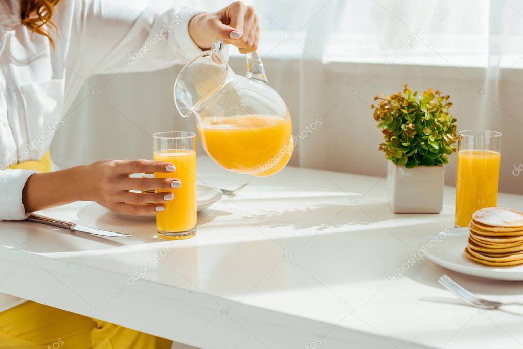 cropped view of woman sitting at table and pouring orange juice in glass while having breakfast