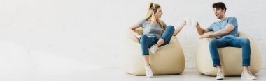 panoramic shot of happy blonde girl and cheerful man holding cups while sitting on bean bag chairs  clipart