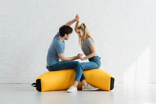 cheerful blonde girl and happy man holding hands while sitting on yellow bean bag chair