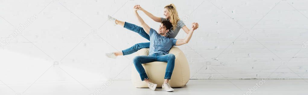 panoramic shot of cheerful blonde girl and happy man holding hands while sitting on bean bag chair 