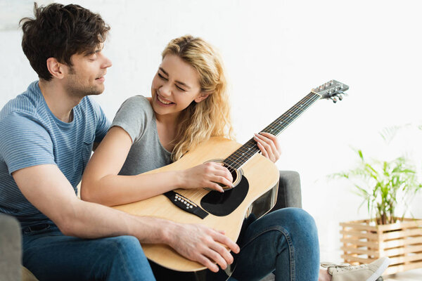 happy blonde woman holding acoustic guitar near cheerful man sitting on sofa 