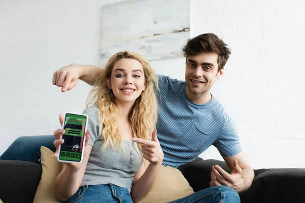 画面上の予約アプリでスマートフォンで指を指差す幸せな男性とブロンドの女性の選択的な焦点 — ストック写真