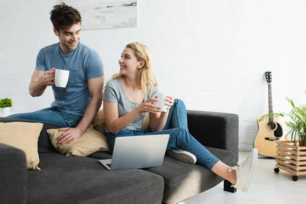 快乐的男人拿着一杯咖啡 坐在坐在几边开朗的金发女孩与笔记本电脑 — 图库照片