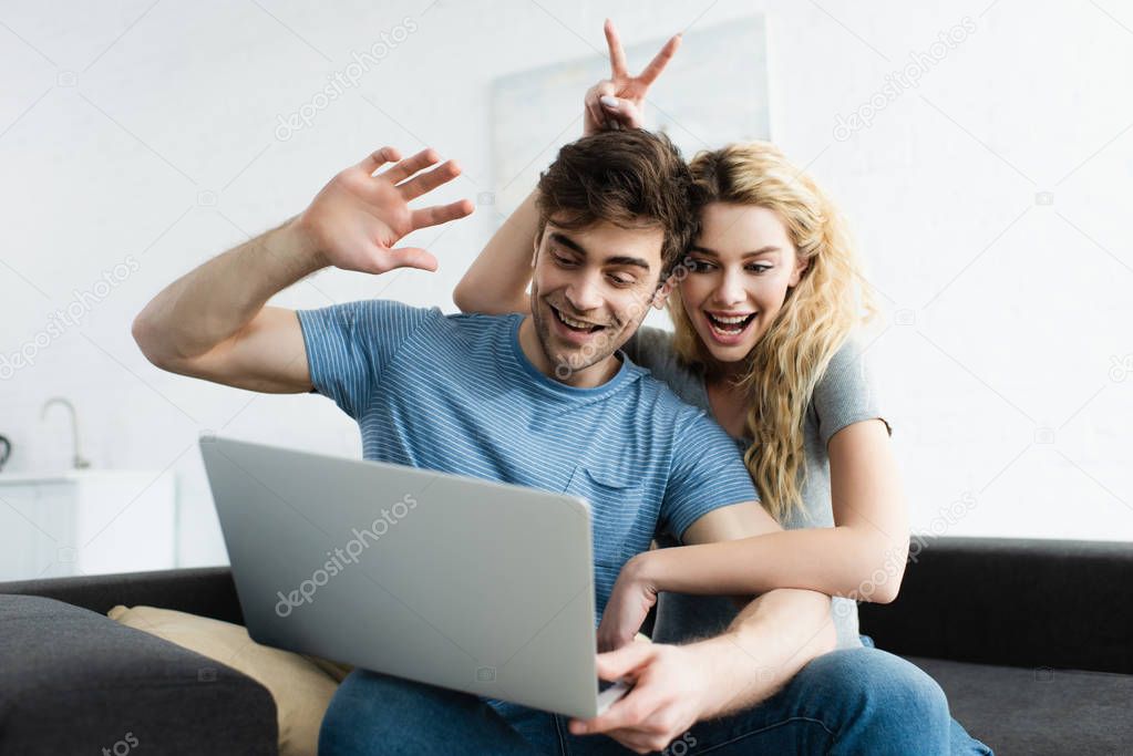 cheerful man waving hand near happy woman showing peace sign while having video call on laptop 
