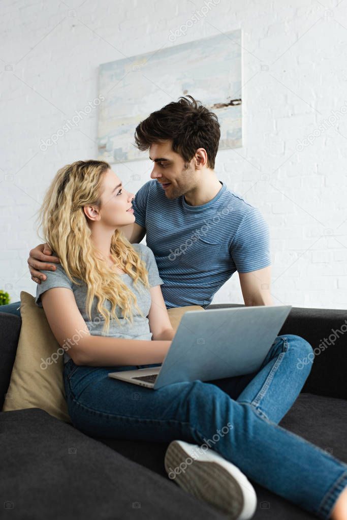 handsome man hugging and looking at happy blonde girl with laptop 