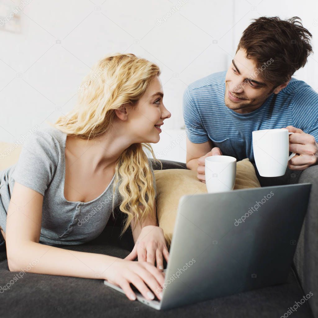 happy and handsome man holding cups near blonde girl with laptop 