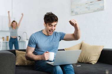 selective focus of happy man celebrating triumph while watching championship on laptop near woman  clipart