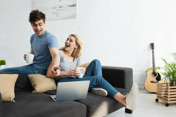 happy man pointing with finger at laptop while holding cup near attractive blonde girl 