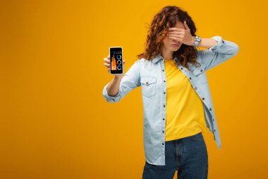 redhead woman covering eyes and holding smartphone with charts and graphs on screen on orange 