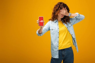 kıvırcık kızıl saçlı kadın gözleri kaplayan ve turuncu ekranda youtube uygulaması ile akıllı telefon tutarak 